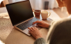 Person typing on a laptop with a cup of coffee on the table, working in a cozy, sunlit environment.